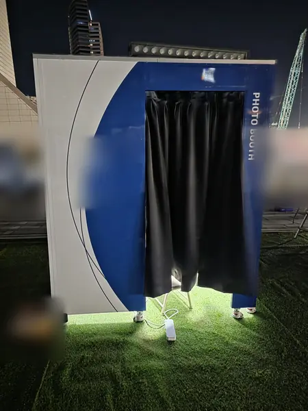 야간 야외 잔디 행사장의 곡선형 파랑·흰색 맞춤 디자인 부스 — 세로 'PHOTO BOOTH' 글자와 검정 커튼