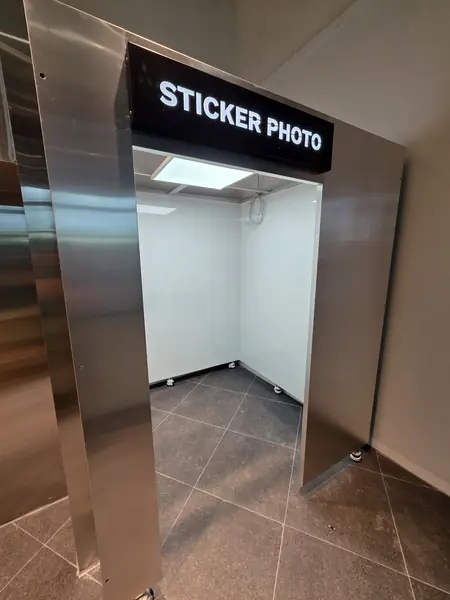 'STICKER PHOTO' 조명 간판을 단 스테인리스 프레임 부스 — 흰 내벽의 열린 촬영 공간