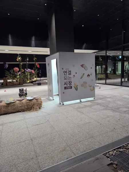 무인양품 「2026 연결되는 시장 · 구황장」 그래픽으로 네 면을 랩핑한 스탠다드 부스 정면 — 야간 실내 광장, 열린 출입구의 흰 조명
