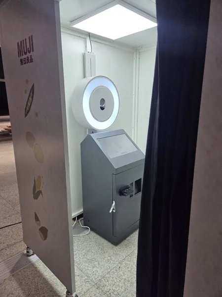 무인양품 랩핑 패널 너머로 보이는 부스 내부 — 천장 조명, 원형 링조명 카메라, 진회색 촬영 키오스크
