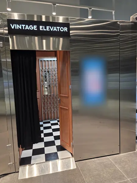 스테인리스 외벽에 'VINTAGE ELEVATOR' 간판이 달린 빈티지 엘리베이터 부스 정면 — 검정 커튼, 원목 문, 흑백 체크 바닥